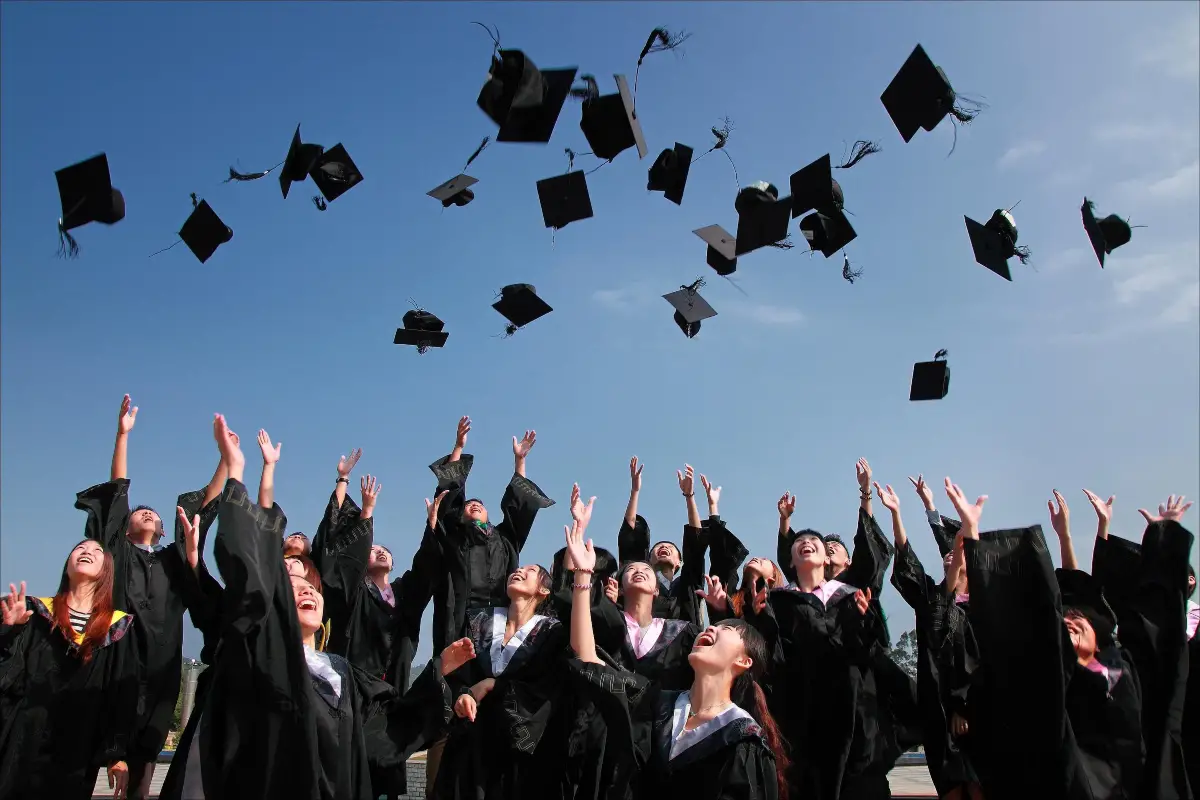 Picture shows a crowd of students wearing graduation gowns, throwing their hats in the air in celebration of graduating from higher education. 