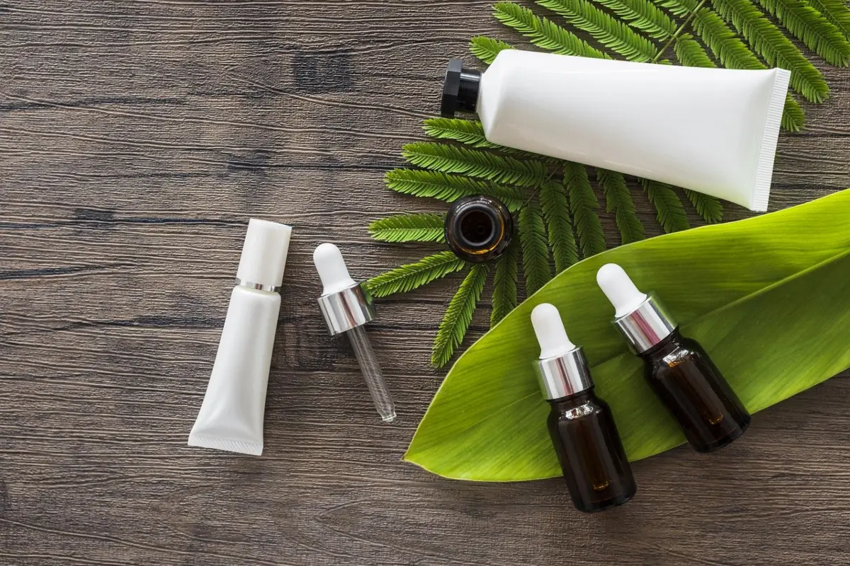 The image shows some plant leaves on a natural wood backdrop wtih a dispensing syringe and four plain (unbranded) bottles of different sizes and shapes suited to various aromatherapy products such as creams, lotions, carrier oils, and essential oils. 