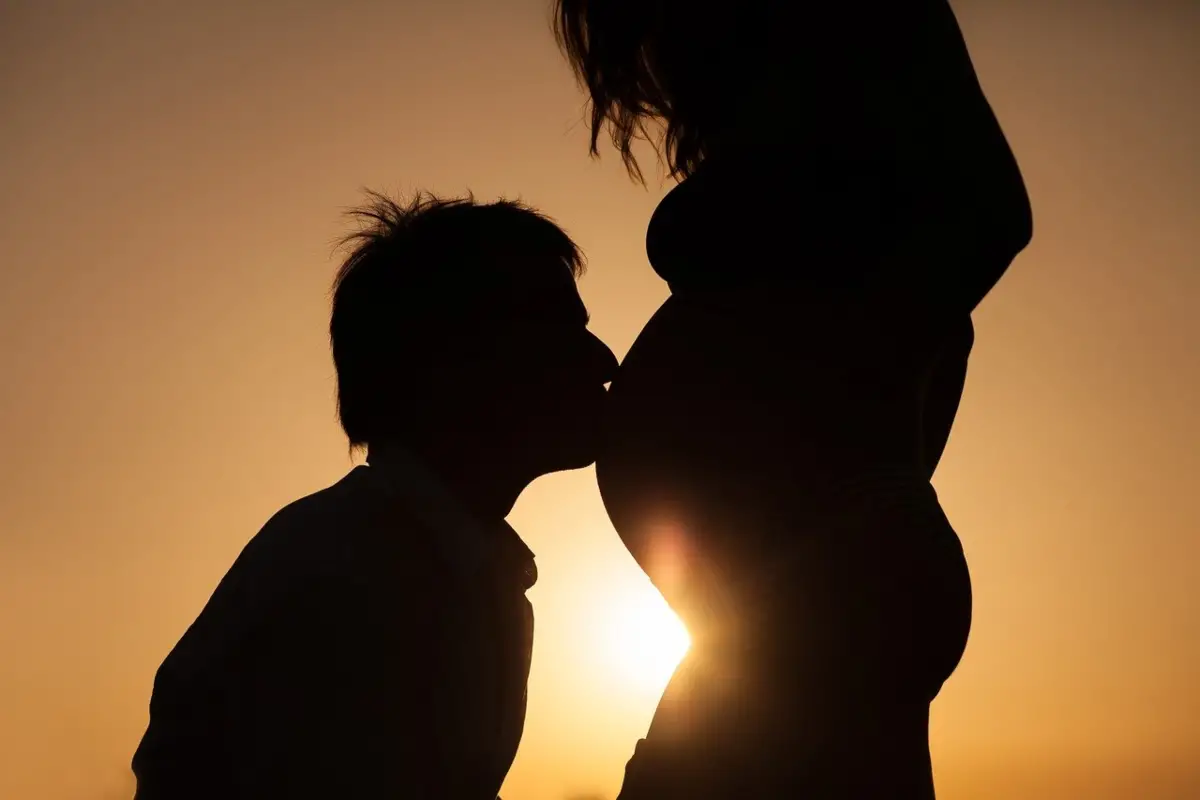 The image is a silhouette image of a couple showing a man kissing the baby bump on his pregnant partner.