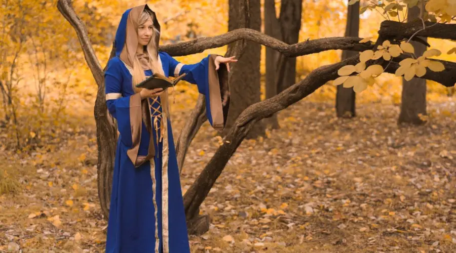 This image shows a young woman dressed in a long Royal blue hooded cloak standing in a forest holding a Book of Shadows which is used for rituals and spell casting. Witchcraft affiliate programs can be used to earn from sales of all witchcraft apparel shown in the picture.